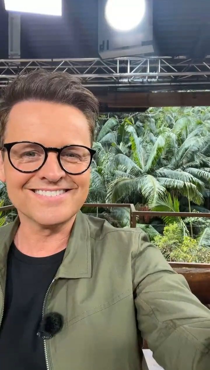 a man wearing glasses and a green jacket is smiling at the camera with palm trees in the background