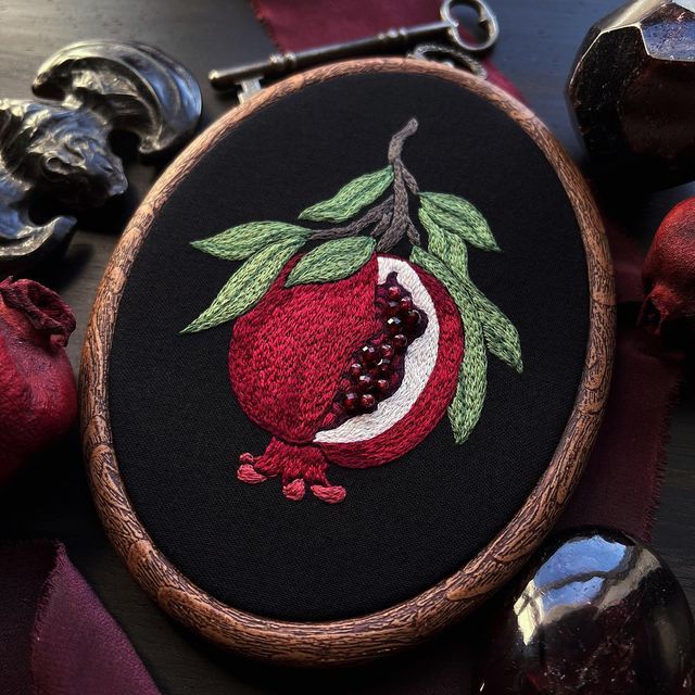 an embroidered pomegranate with leaves and berries on black fabric surrounded by other items