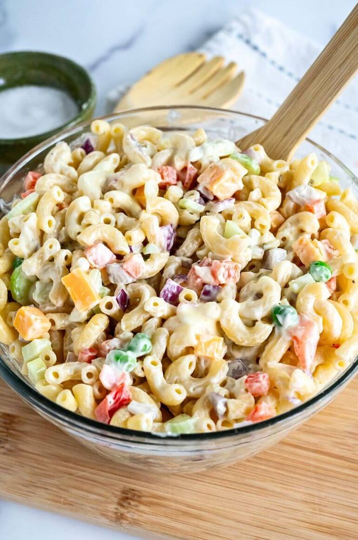 a glass bowl filled with macaroni salad on top of a wooden cutting board