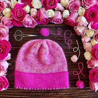 a pink knitted hat surrounded by flowers and knitting needles on a wooden table with scissors