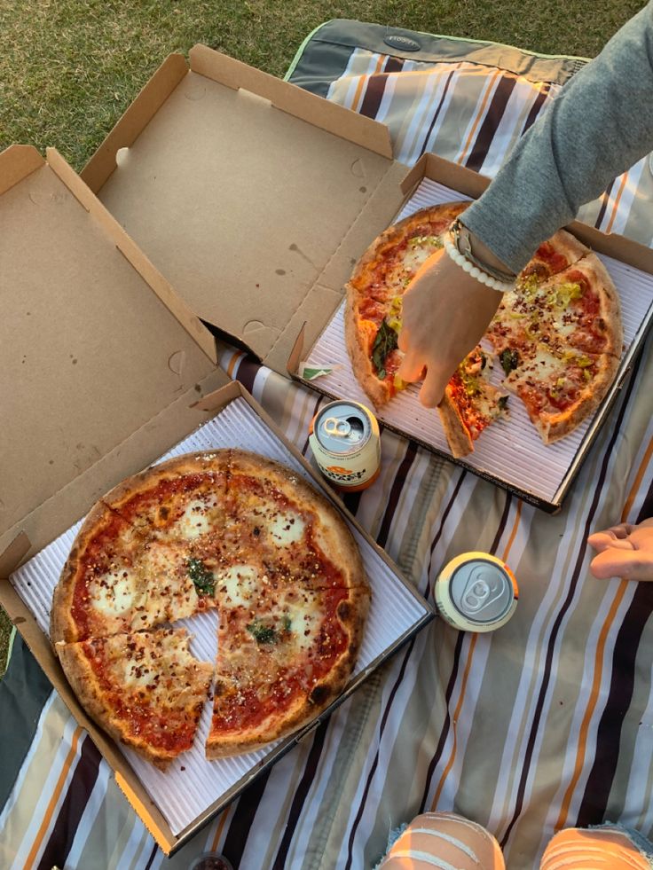 two pizzas sitting on top of a picnic blanket next to cans of soda and water