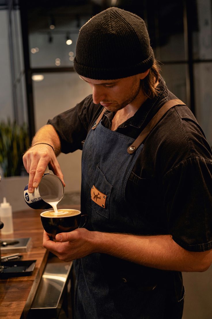 a man pours coffee into a cup in his kitchen while wearing an apron and hat