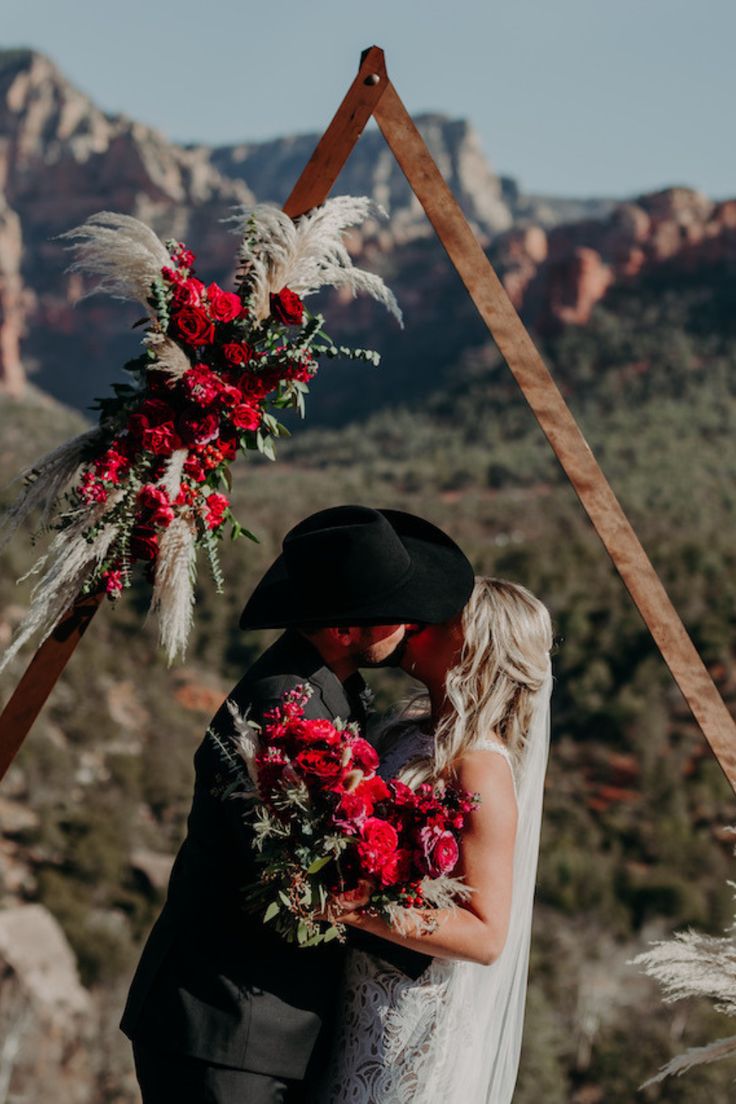 a bride and groom are standing under an arch decorated with red flowers in the mountains