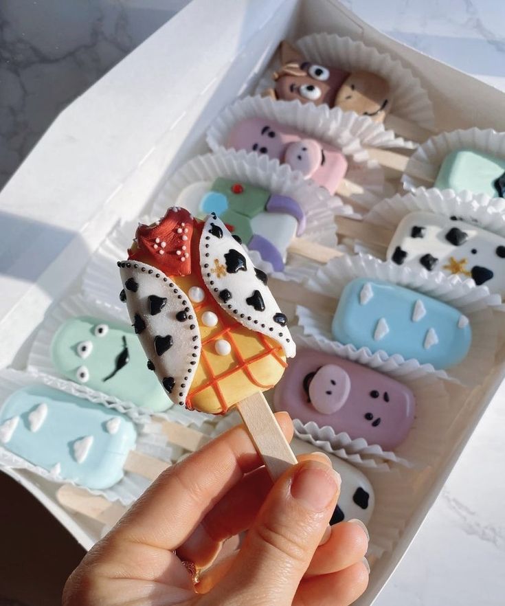 a hand holding a toothpick in front of several decorated cupcakes