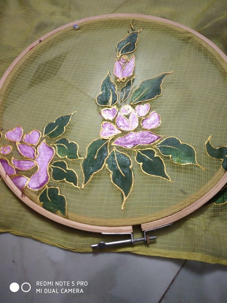 an embroidery project with pink flowers on green fabric and gold trim around the edges, sitting on top of a piece of cloth