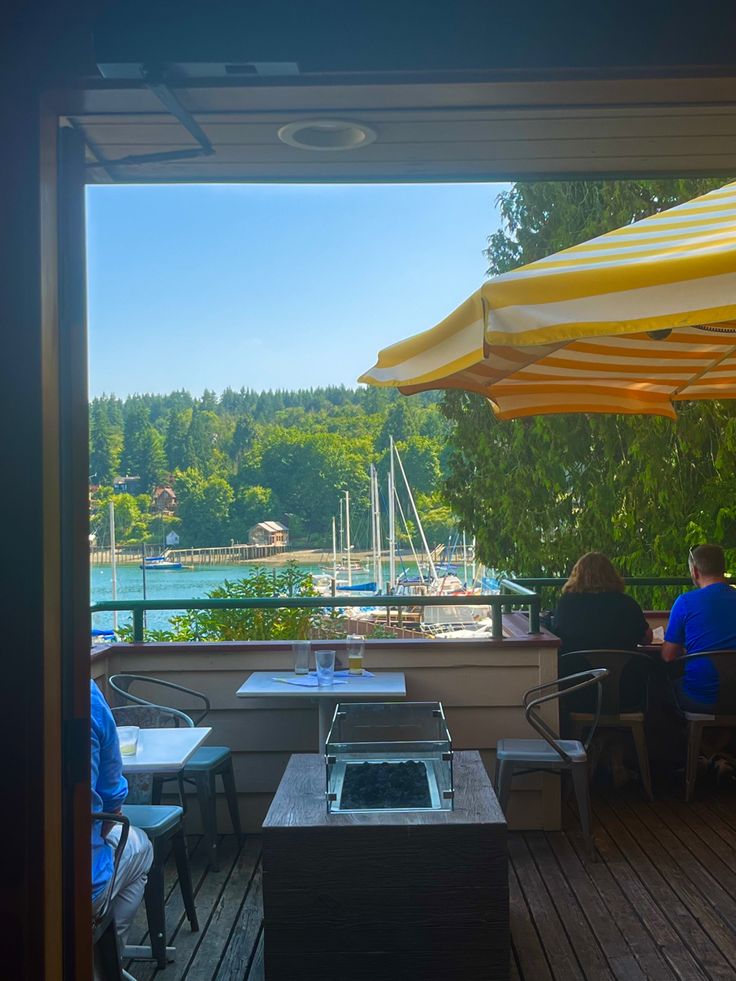 two people sitting at an outdoor table with umbrellas over them and boats in the water