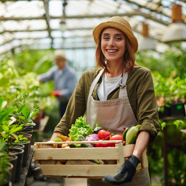 a woman holding a crate full of vegetables in a greenhouse