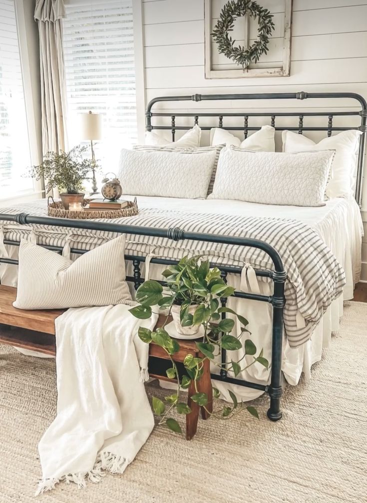 a bed with white sheets and pillows in a bedroom next to a window filled with potted plants