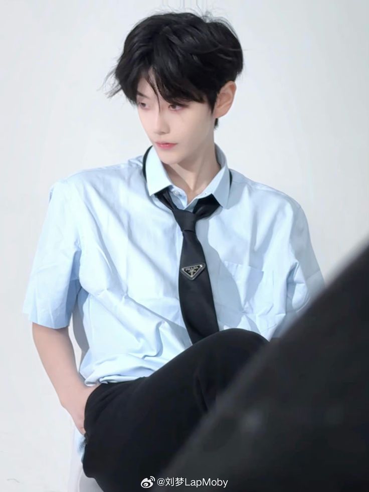 a young man wearing a blue shirt and black tie sitting on a white chair with his hands in his pockets