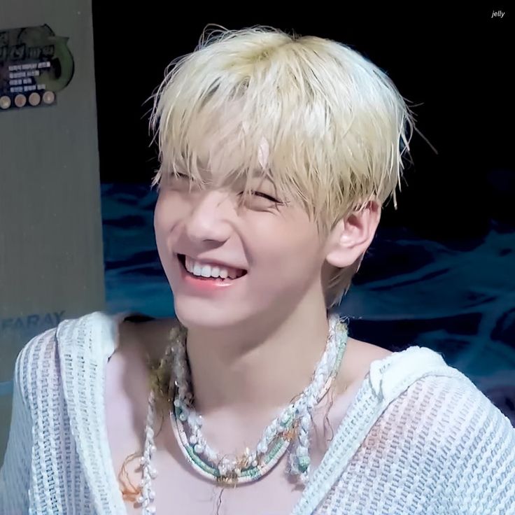 a young man with blonde hair smiling and wearing a pearl necklace in front of a black background