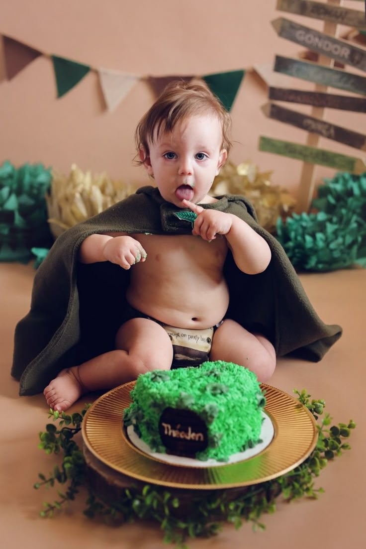 a baby sitting in front of a cake with a cape on it's head