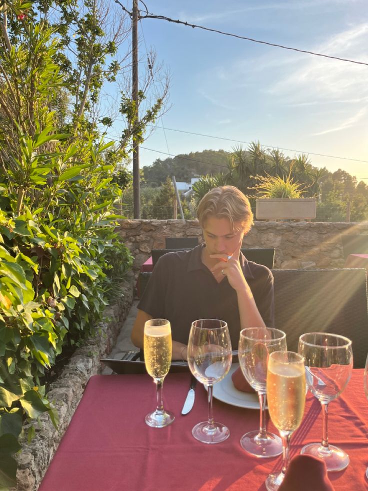 a woman sitting at an outdoor table with wine glasses on it, and another person in the background