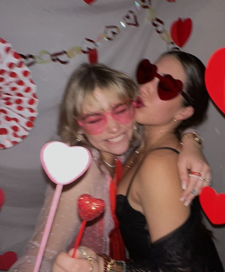 two women hugging each other while holding lollipops in front of a heart - shaped backdrop