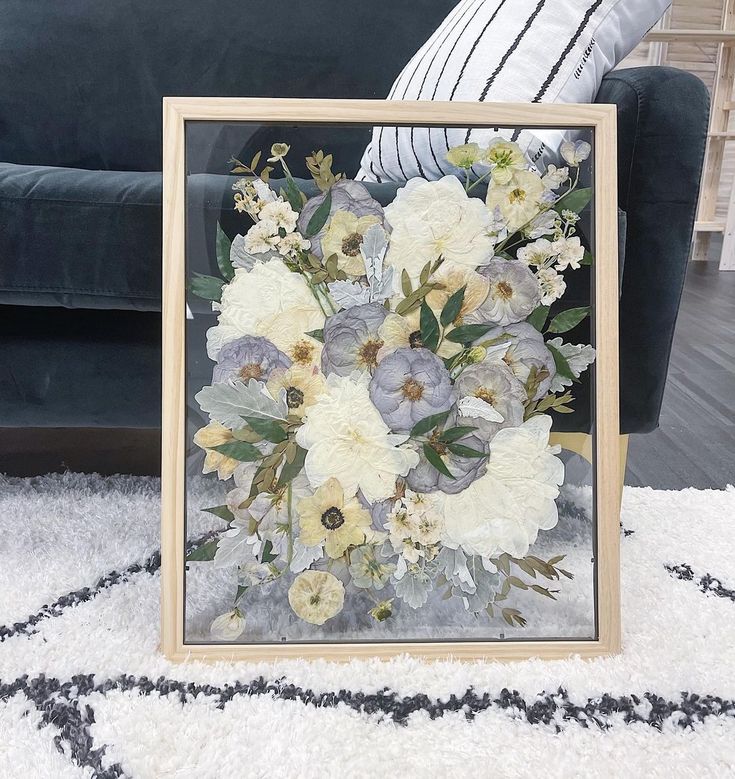 an arrangement of white and grey flowers in a wooden frame on a rug next to a couch