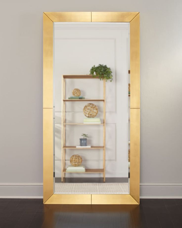 a gold framed mirror on the wall with shelves and plants in it, against a white wall