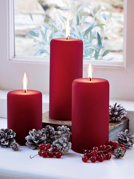 three red candles sitting on top of a window sill next to pine cones and berries