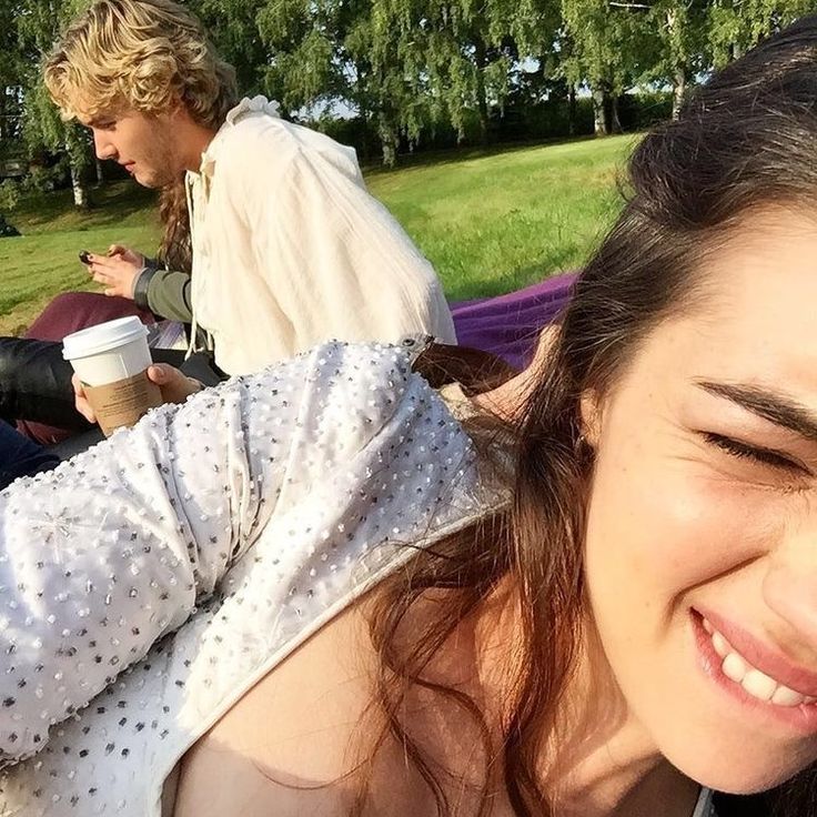 a woman is smiling while sitting in the grass with a man behind her on a cell phone