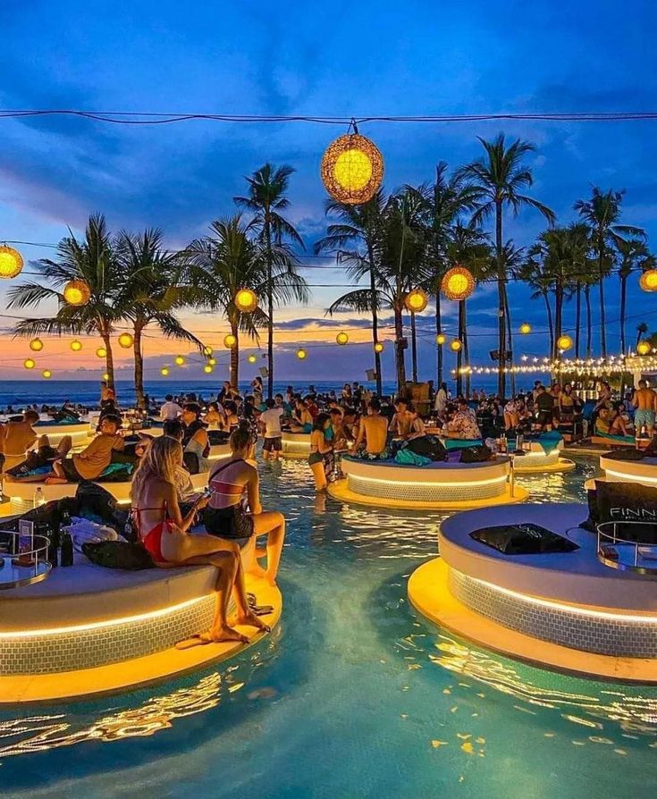 many people are sitting at tables near the water and some have lights hanging above them