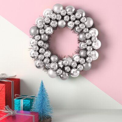 a christmas wreath with silver balls and tinsel on it, next to presents in front of a pink wall