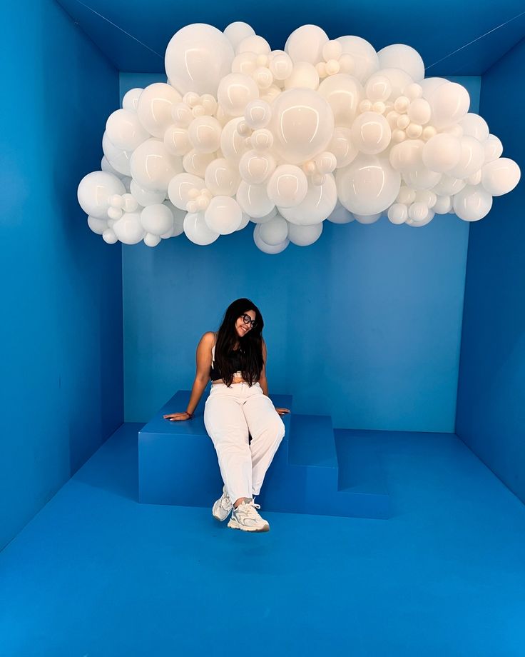 a woman sitting in a blue room with white balloons floating above her and on the floor