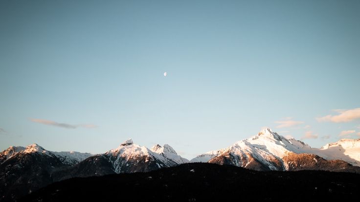 the mountains are covered in snow as the sun is low to the ground and there is a half moon above them