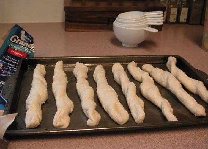 bread sticks are lined up on a baking sheet with white bowls and utensils in the background