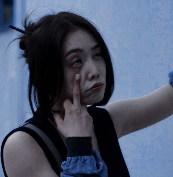 a woman holding her hand up to her face and looking at the camera while standing in front of a blue wall