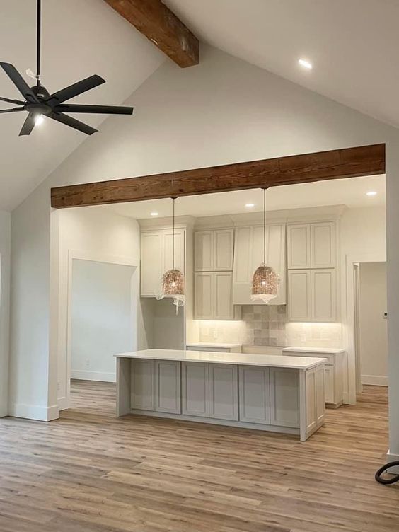 an empty kitchen and living room in a house with wood floors, ceiling fan and white cabinets