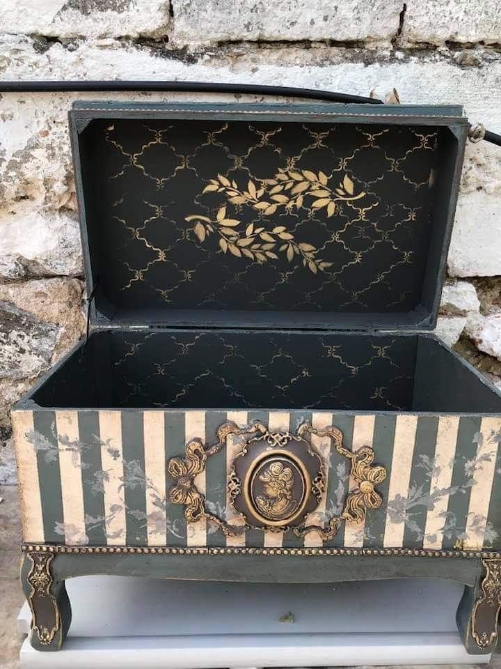 an old trunk is painted black and white with gold leaf designs on the lid, sitting against a stone wall