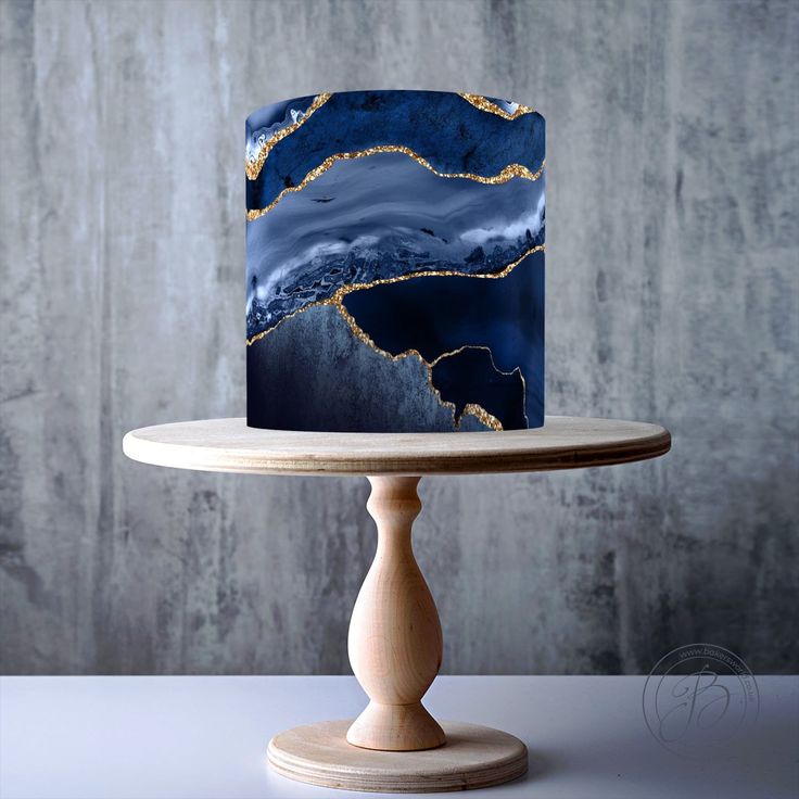 a blue and gold marbled cake on a wooden stand against a gray wall with the top layer partially covered in icing