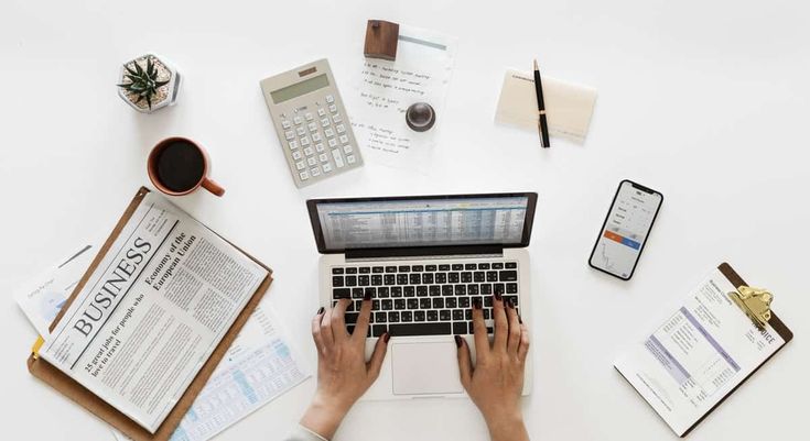 a person typing on a laptop computer surrounded by papers and calculator, coffee mugs