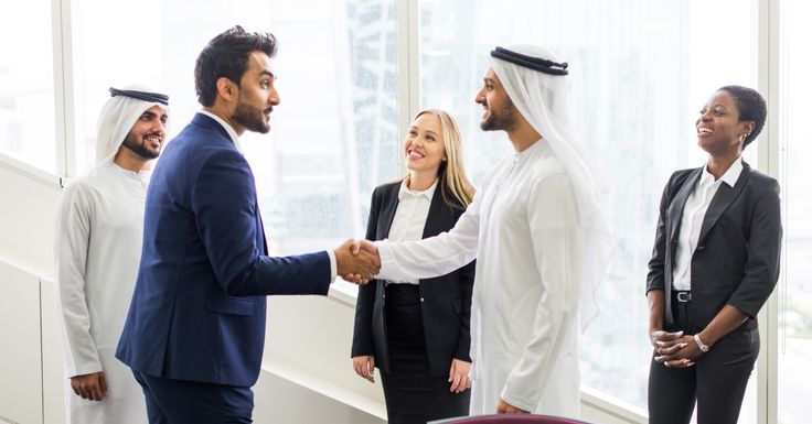 two men shaking hands with three other people in front of a window, one wearing a headscarf