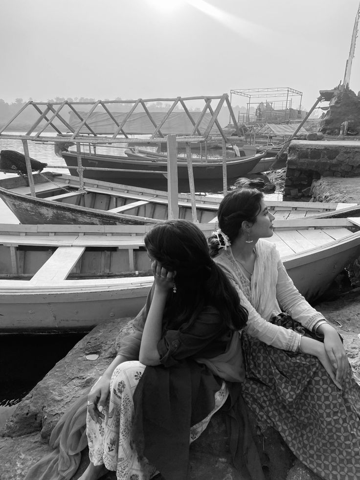 two women sitting on the edge of a pier next to boats