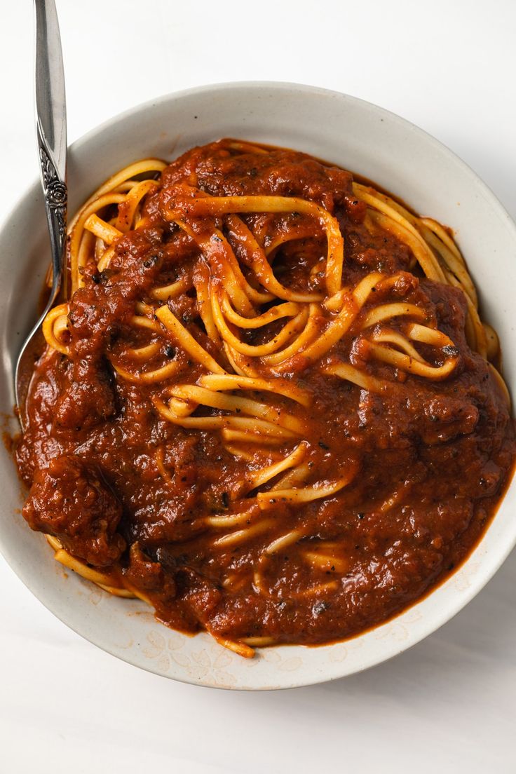 a white bowl filled with spaghetti and sauce on top of a table next to a fork