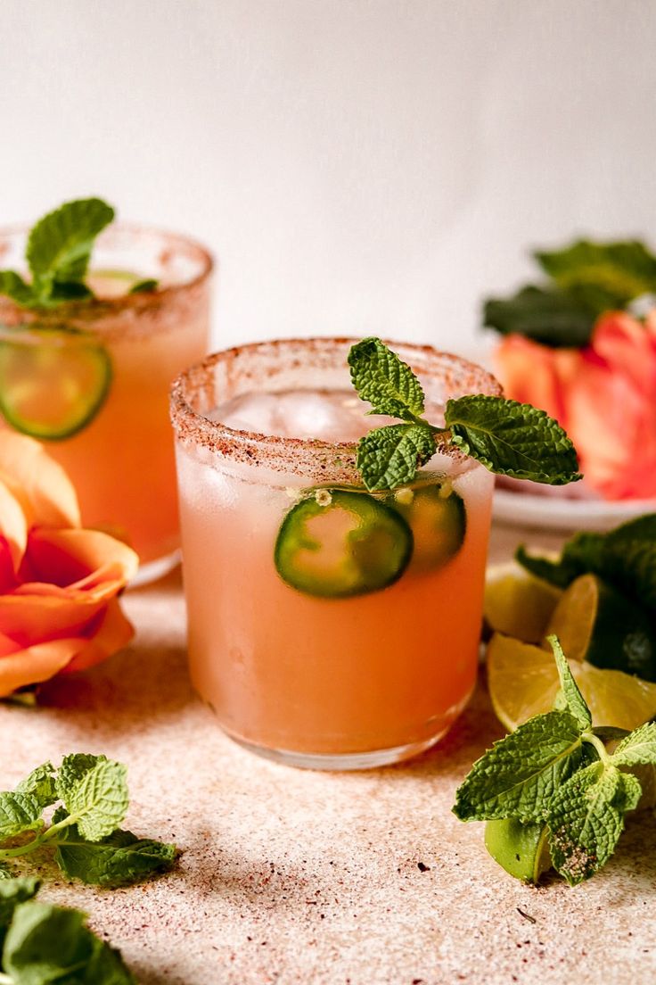 two glasses with cucumber and mint garnish sit on a table next to flowers