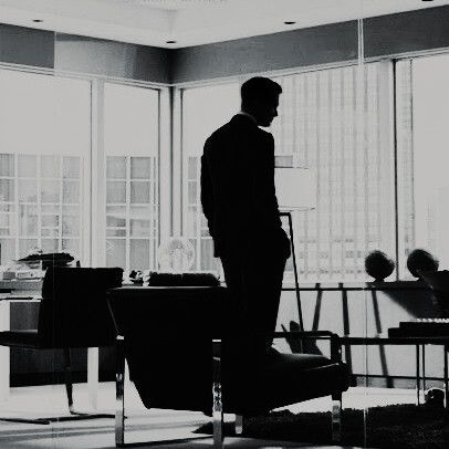 a man standing in an office looking out the window at another person sitting on a chair