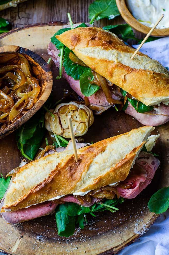 a sandwich with meat, onions and greens on a wooden platter next to bread