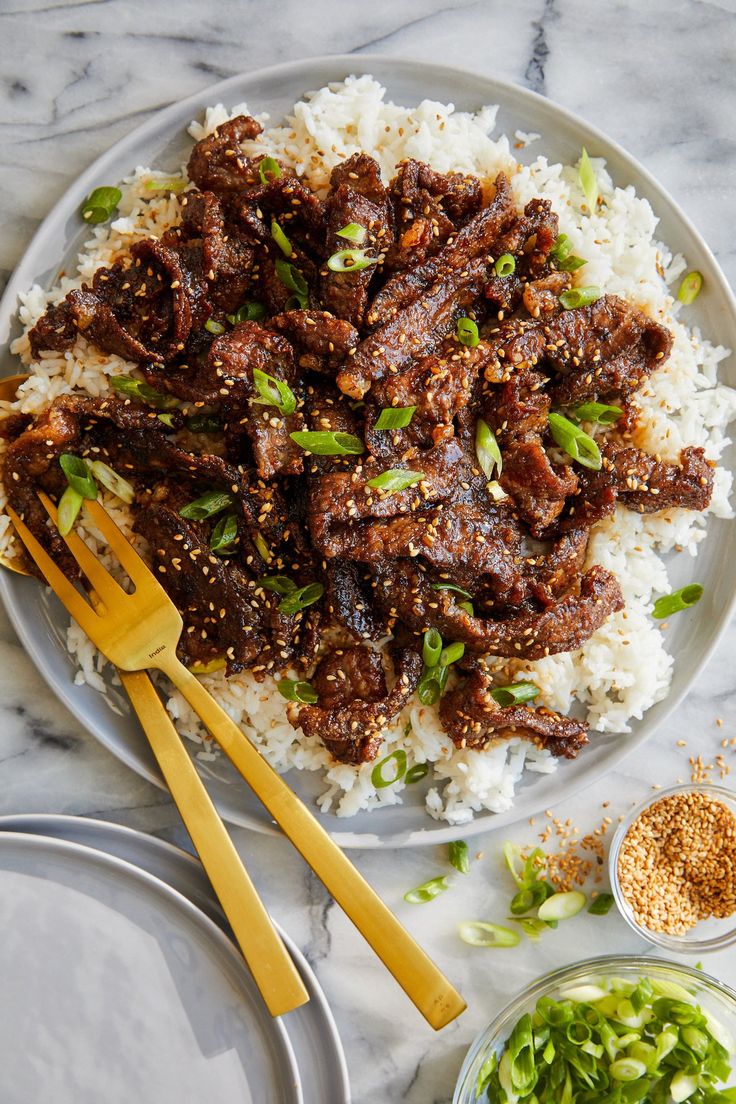 a white plate topped with meat and rice next to two bowls of green vegetables on a marble table