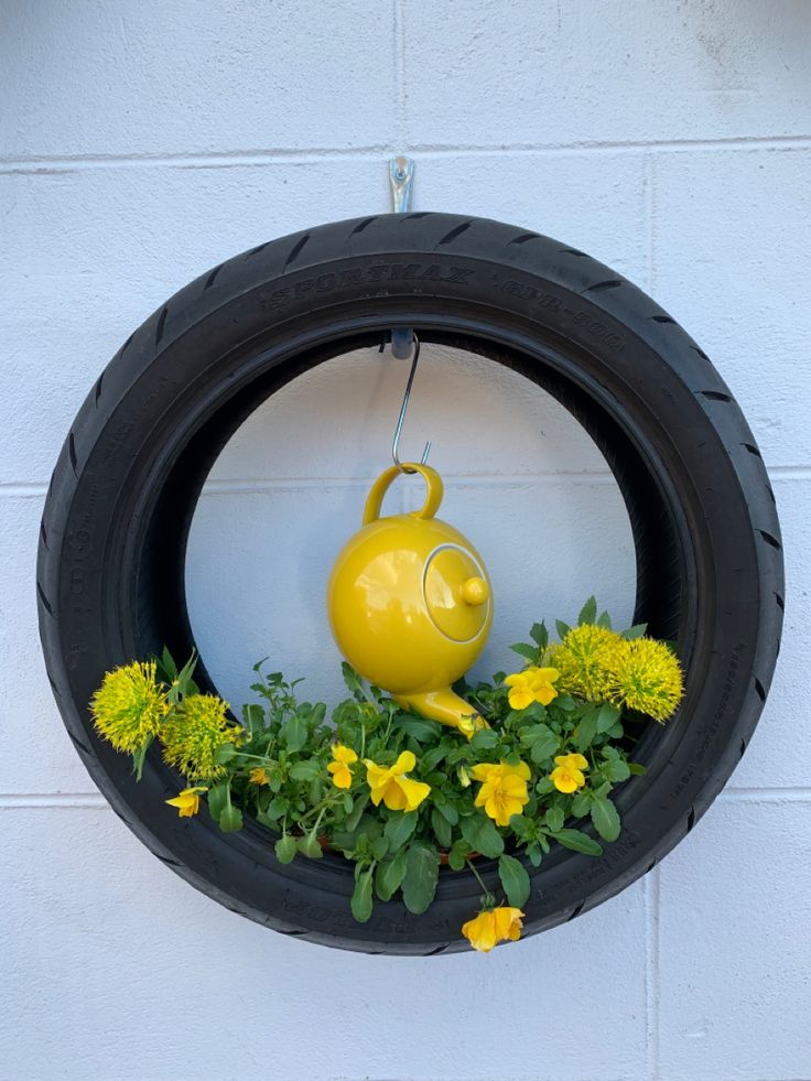 a yellow teapot in a tire planter filled with green and yellow flowers on the side of a white brick wall