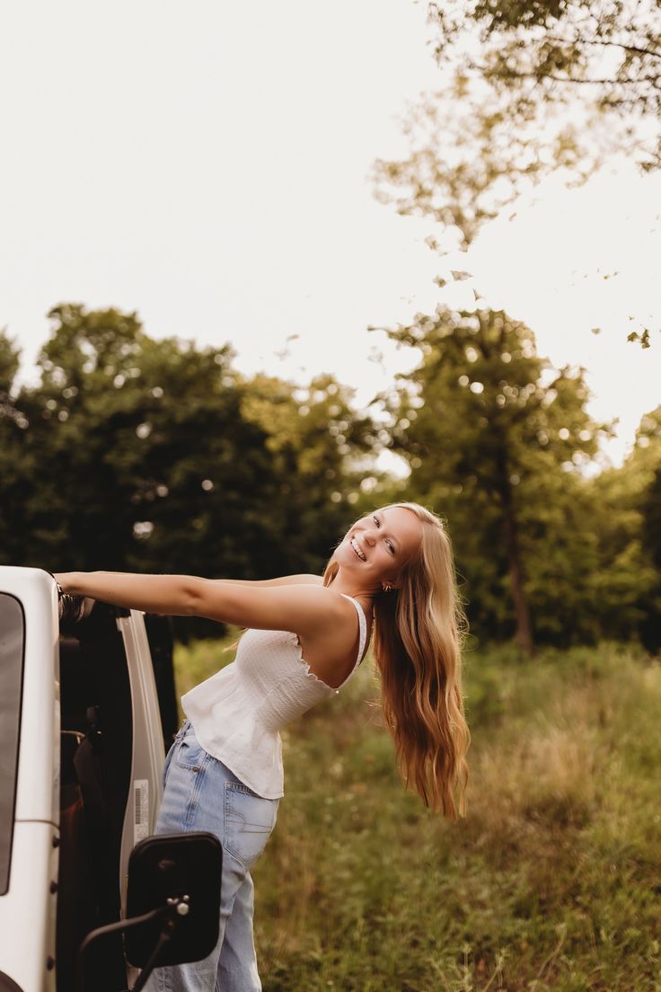 a woman leaning on the back of a white truck in a field with trees behind her