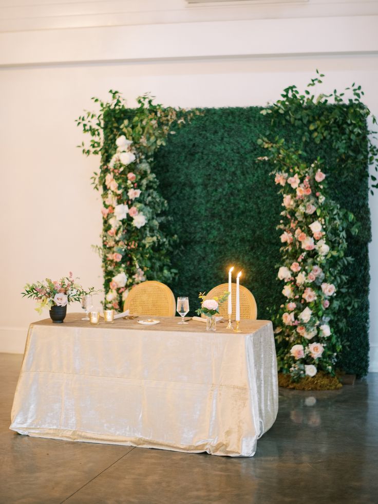 a table with candles and flowers on it in front of a green wall that is decorated with greenery