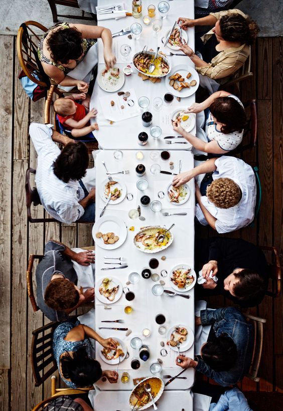 a group of people sitting around a long table eating food and drinking wine at it