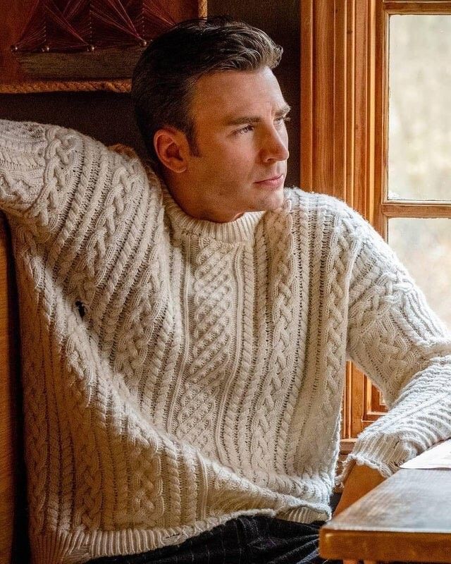 a man sitting at a table in front of a window wearing a sweater and jeans