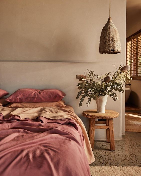 a bed with pink sheets and pillows next to a wooden stool in front of a window