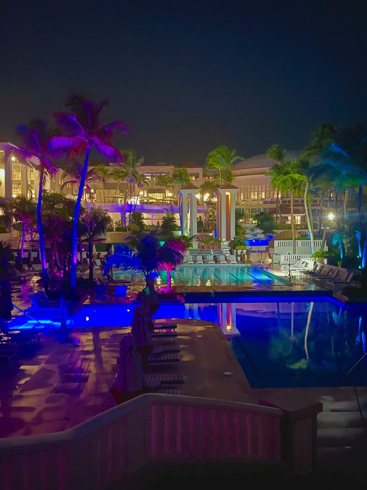 an outdoor swimming pool at night with palm trees and buildings in the background as well