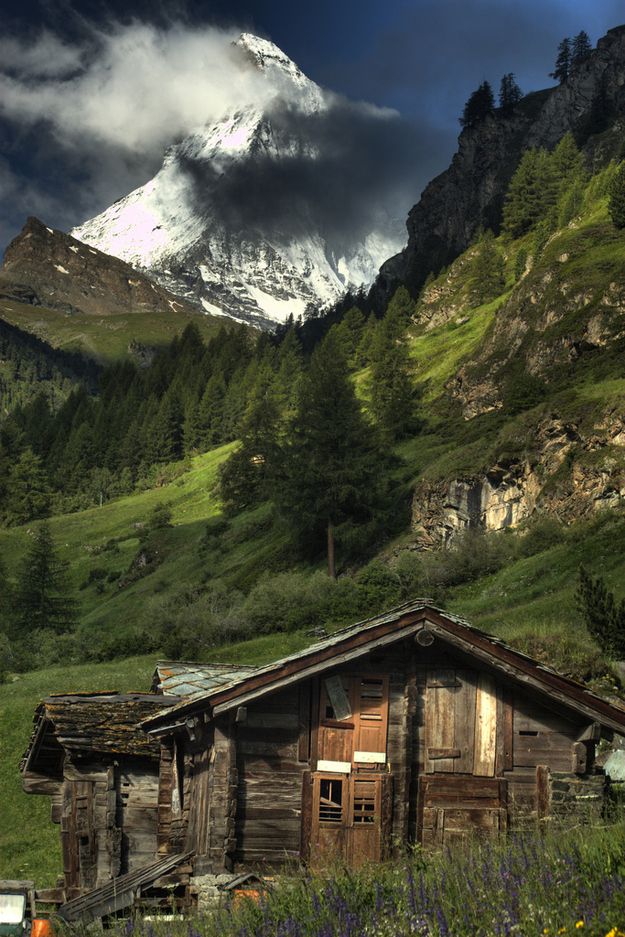 an old wooden cabin in the mountains with snow on the mountain behind it and green grass below