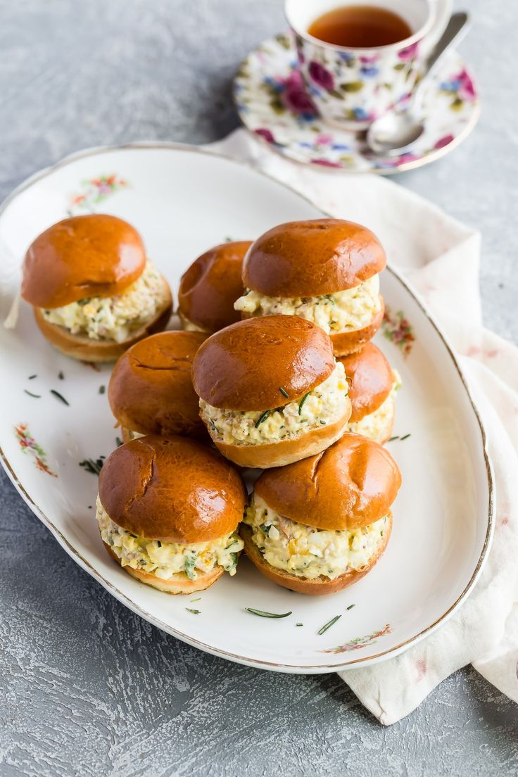 several small sandwiches on a plate with a cup of tea in the backgroun