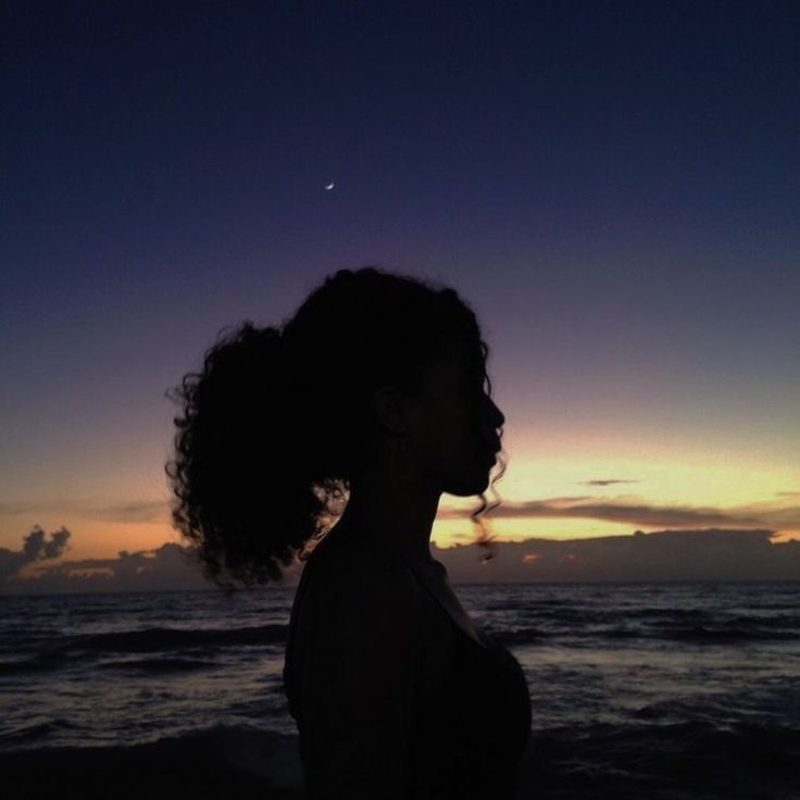 a woman standing on top of a beach next to the ocean at sunset with her hair blowing in the wind