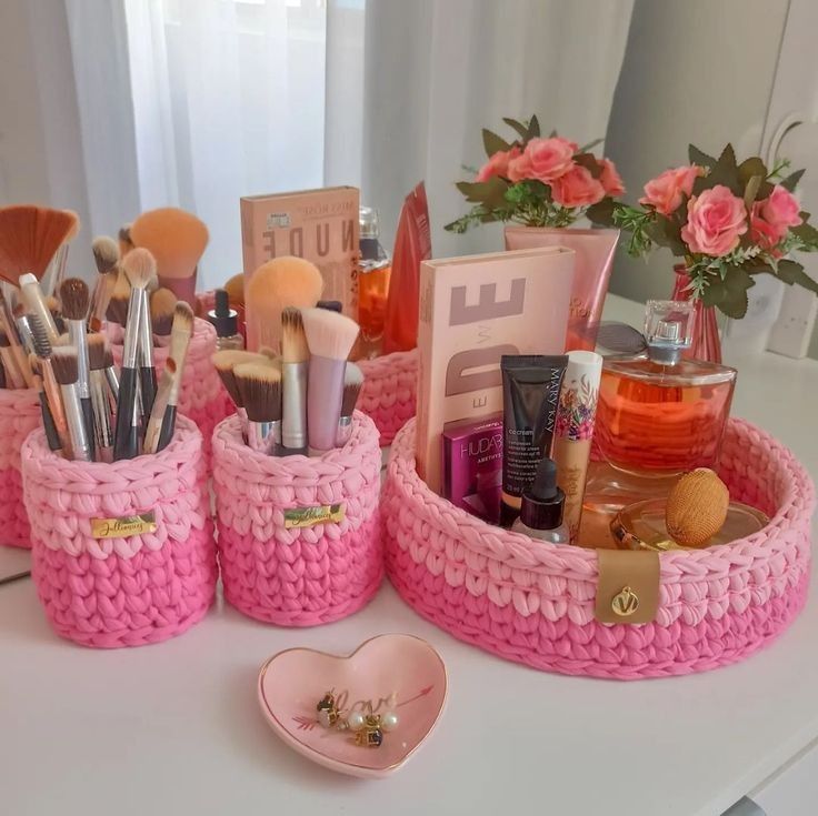 pink crocheted containers with makeup and other items in them on a white table