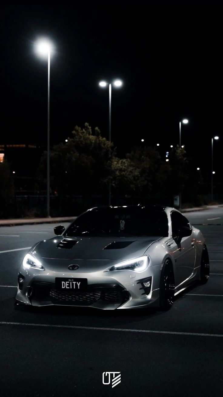 a silver sports car parked in a parking lot at night with street lights behind it